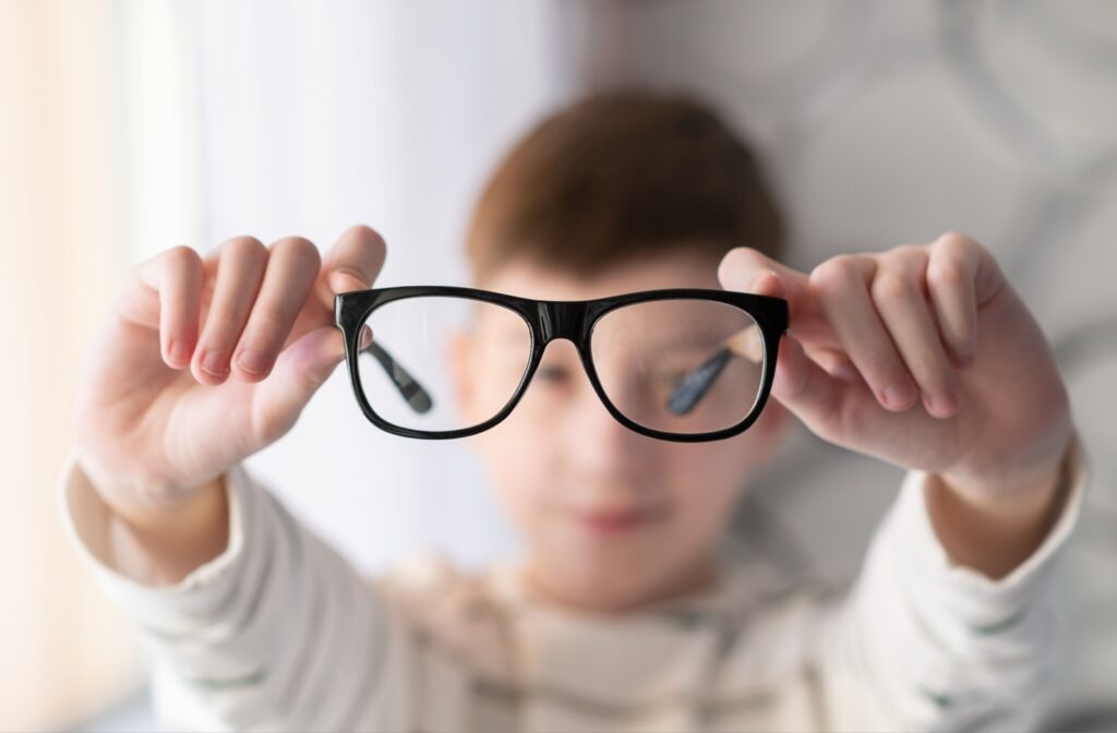A child is holding a pair of black-framed glasses up to the camera, with their face blurred in the background.