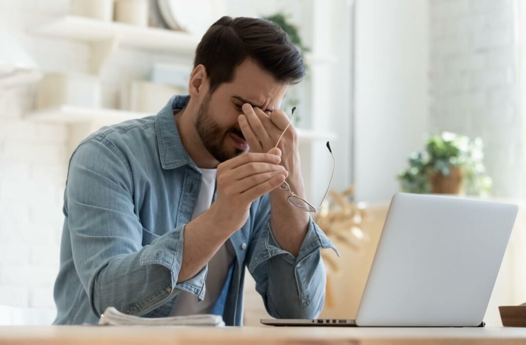 A person wearing a denim shirt sits at a laptop and removes their glasses to rub their eyes.