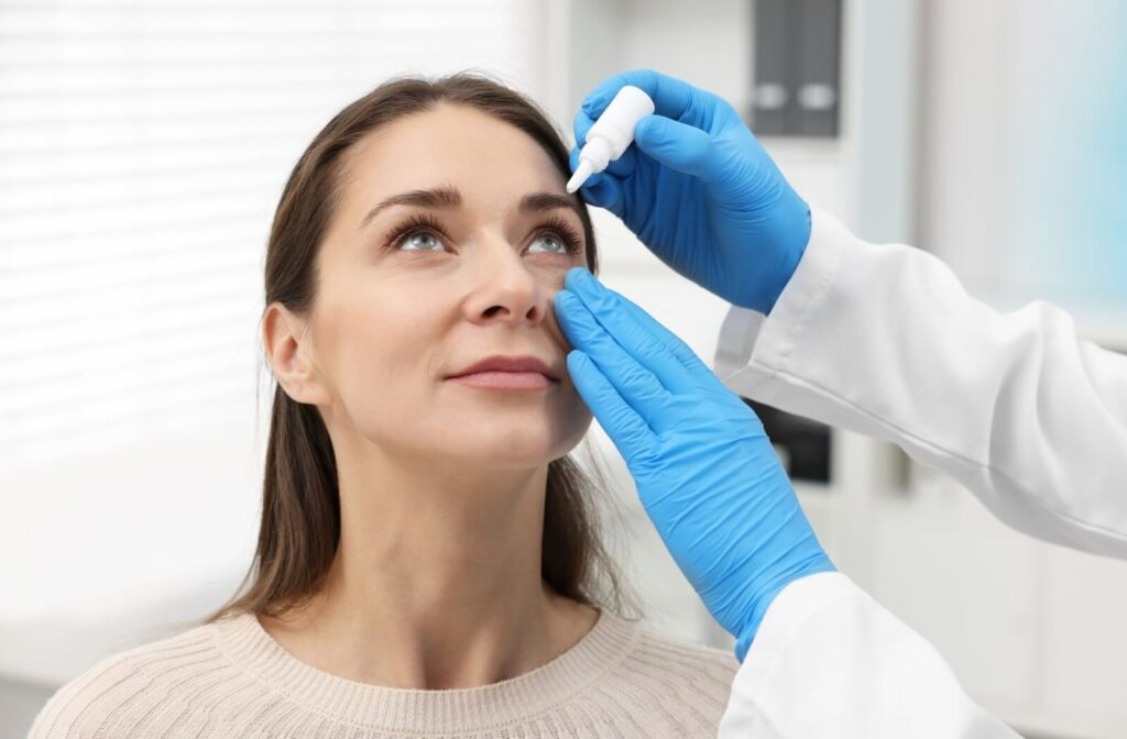 An optometrist assists a patient with applying eye drops by gently pulling down her eyelid and using a bottle.
