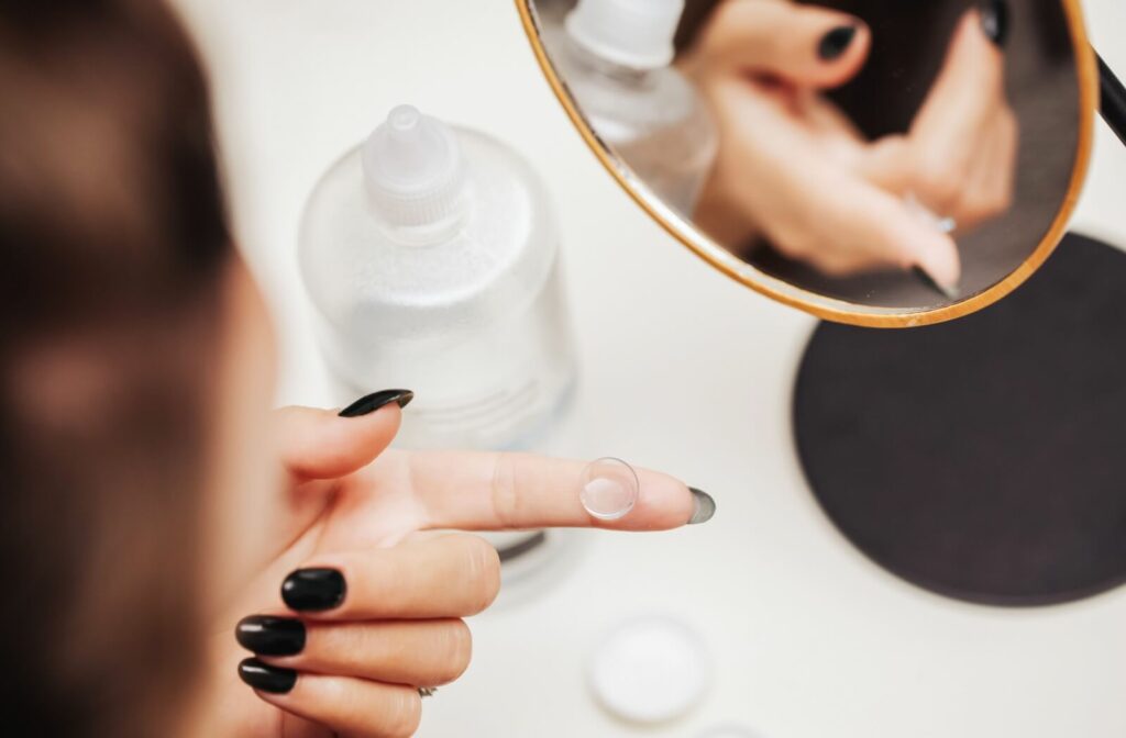 A woman with black nail polish holding a clear contact lens on her fingertip, ready for insertion in front of a mirror with saline solution nearby.