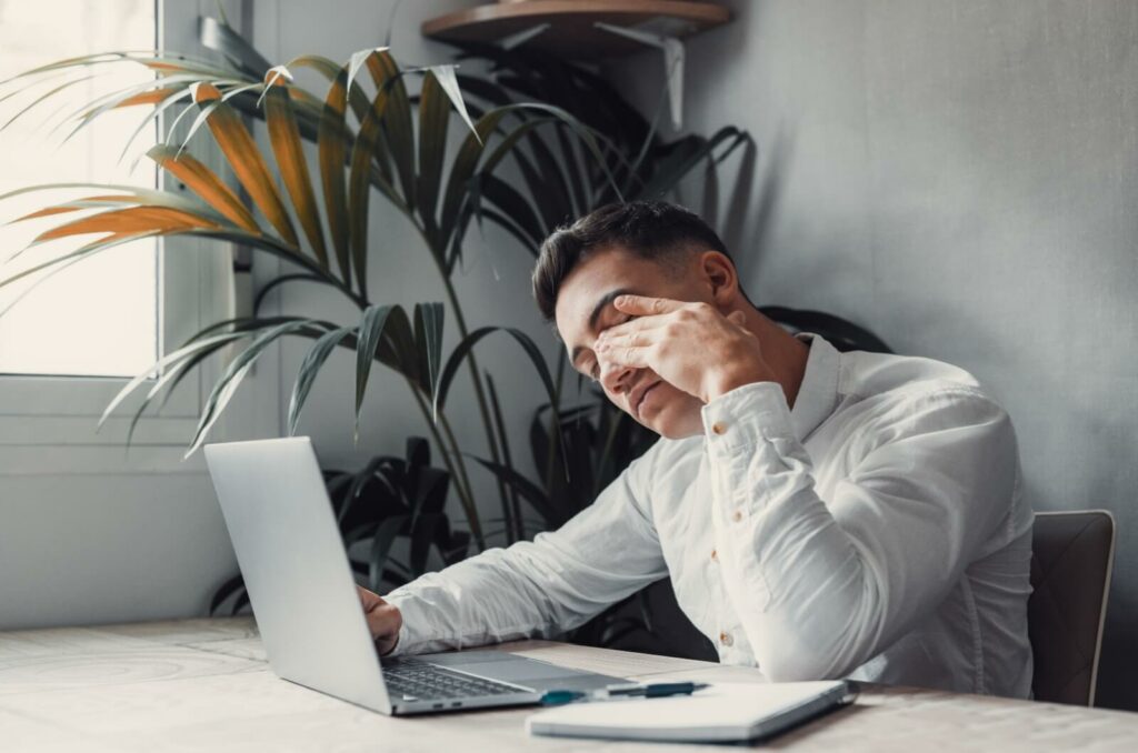 Man sitting at a desk with a laptop, rubbing his eyes from strain while working.