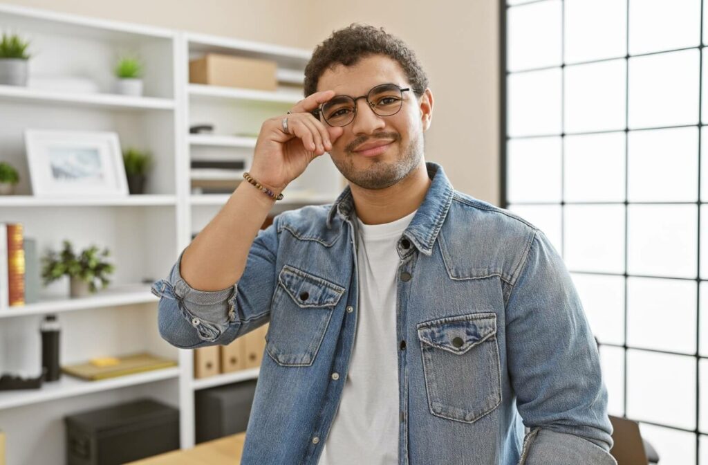 A smiling person in a modern office adjusts their glasses.