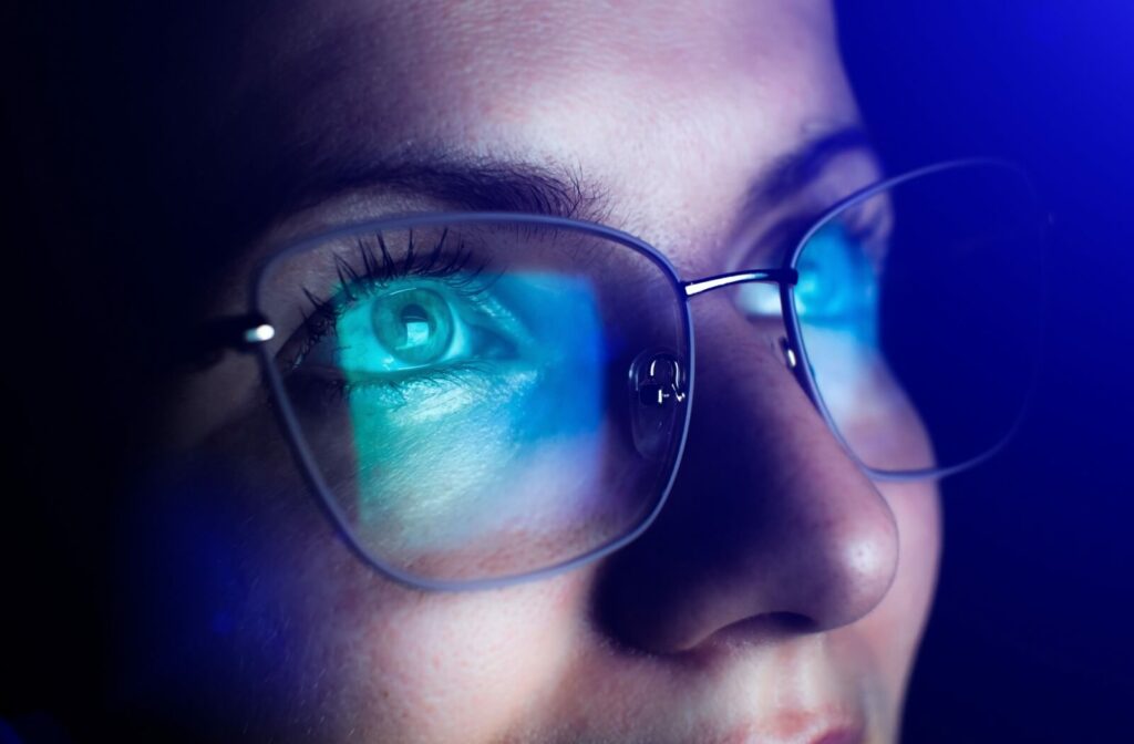 A close-up of a person wearing blue light glasses in a dark room. The light of a digital screen reflects off the lenses.