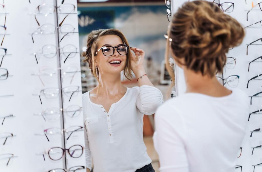 A woman smiling and trying on a pair of eyeglasses while looking in a mirror at an optical store