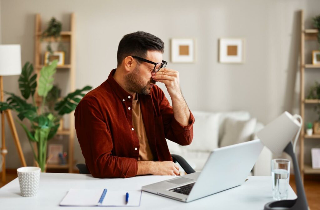 A person wearing glasses sits at a desk with a laptop, rubbing the bridge of his nose from eye strain or discomfort.