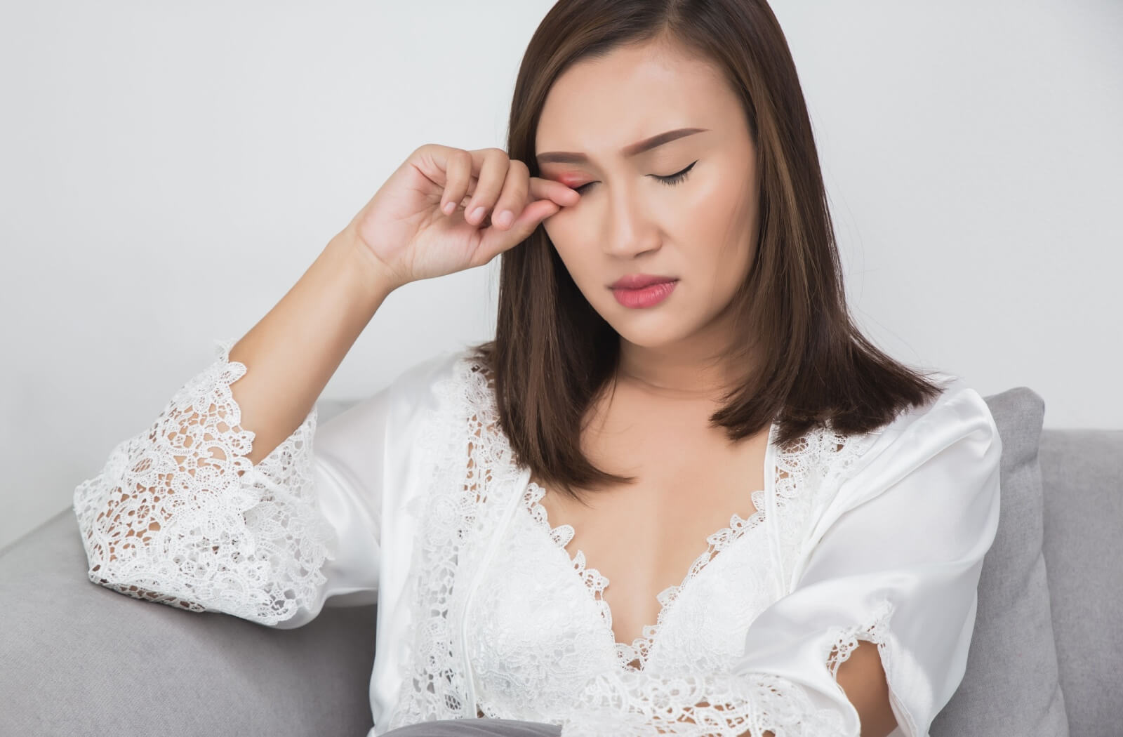 A person sitting on a sofa in a white lace robe, eyes closed and gently rubbing an eyelid, demonstrating a common symptom of clogged Meibomian glands.