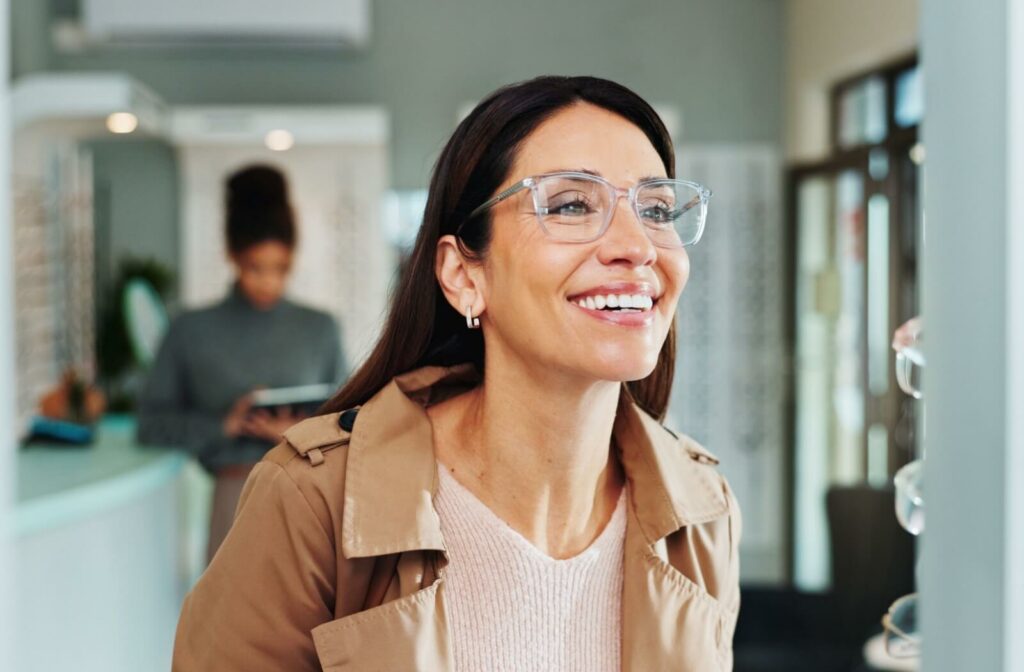 A person wearing clear-rimmed glasses smiling while looking in a mirror at an optical shop.