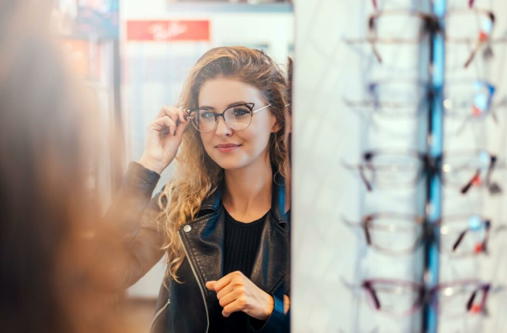A person with curly hair wearing a leather jacket and dark cat-eye glasses looking into a mirror.